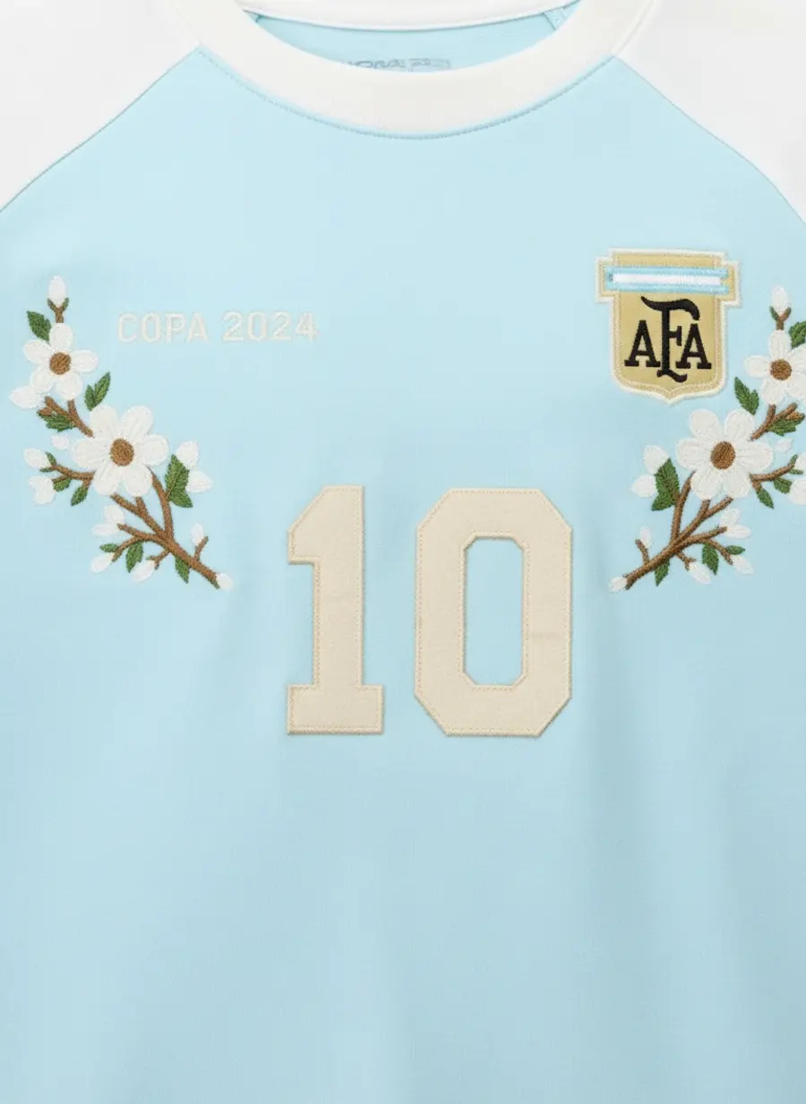 Close up of embroidered flowers, a woven club badge and a large appliqué number on a pale blue raglan football tee