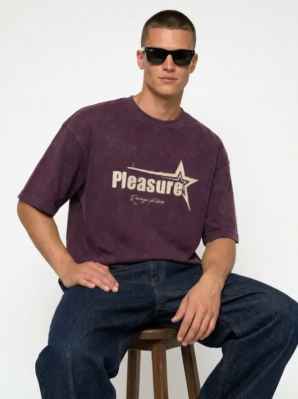 Washed plum Pleasure star-graphic tee with indigo denim, seated on a stool, on an AI male model