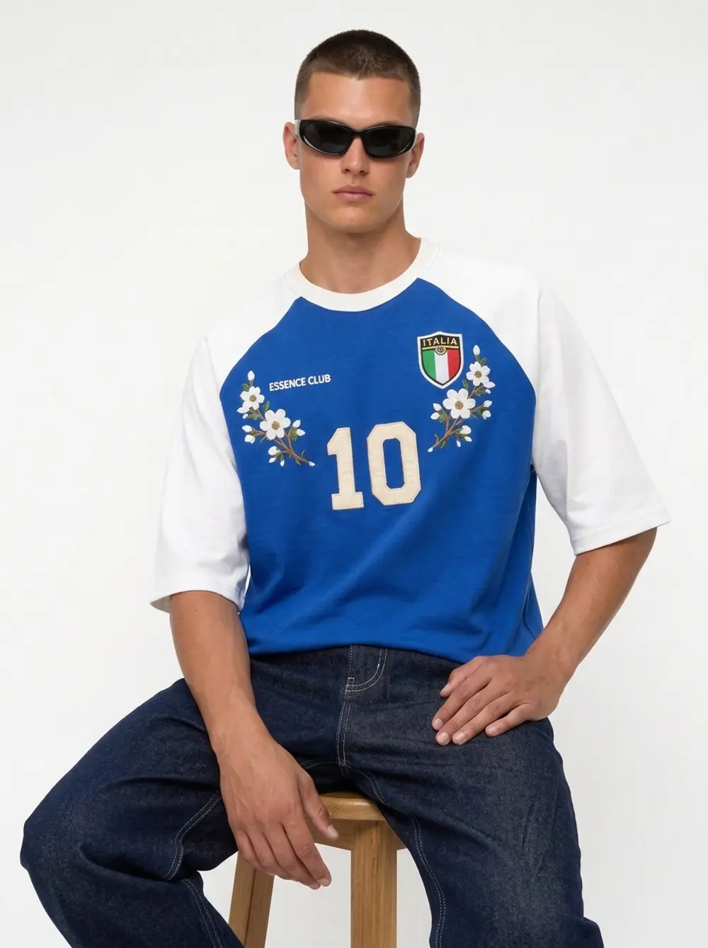 Blue Essence Club raglan football tee, seated on a stool in black sunglasses, on an AI male model