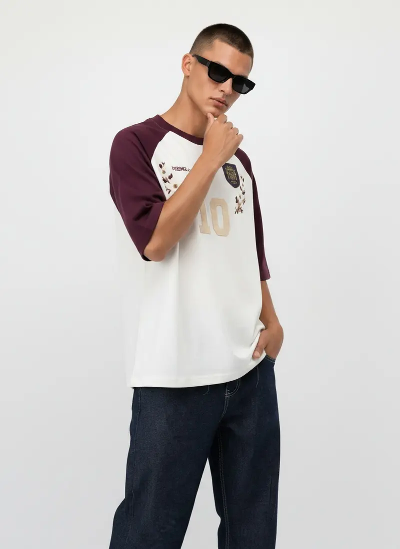 Bully Essence Club Raglan in burgundy / cream on the same male model in black sunglasses in a white studio, posed differently from the other colourways