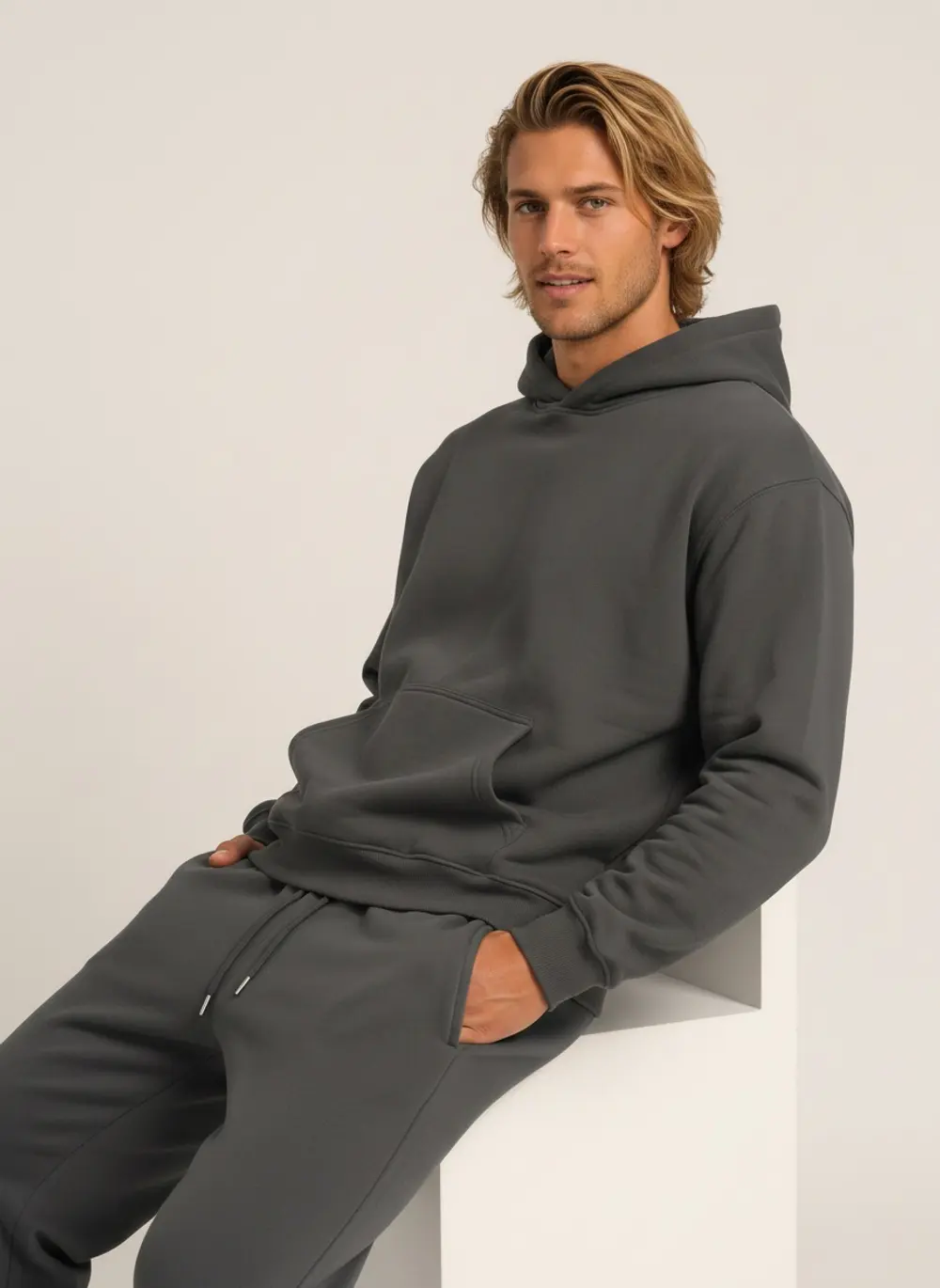 Graphite hooded sweatshirt and jogger set, seated on a block, on an AI male model in a warm studio