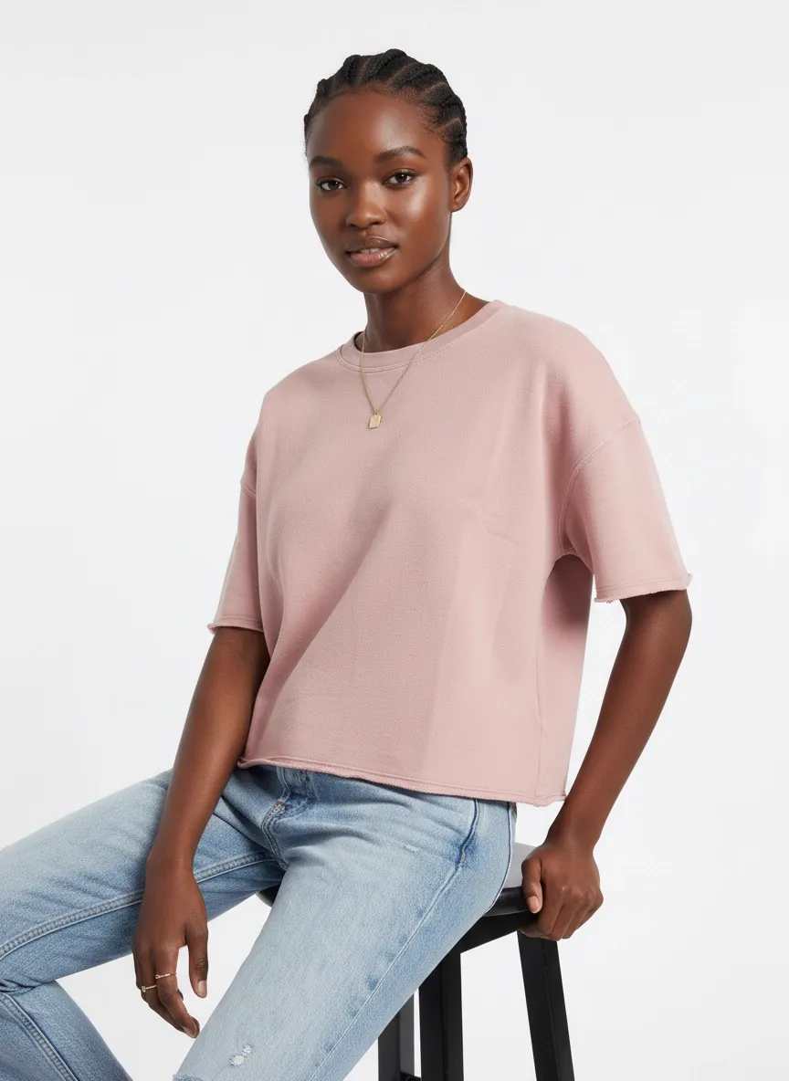 MODA AI catalog output — Black model in pink boxy tee leaning against a stool, editorial PDP styling with light denim and a leather bag