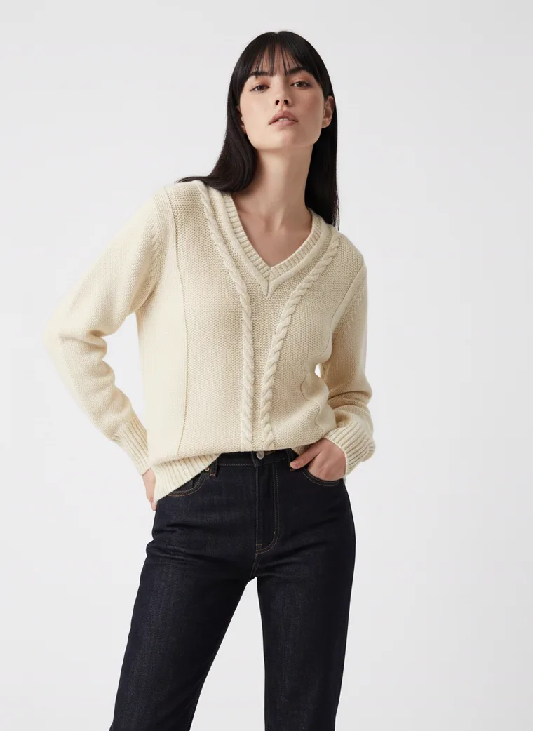 A cream cable knit v-neck jumper with black straight jeans on pale grey. Cable Knit V-Neck in Cream, frame 1 of 5