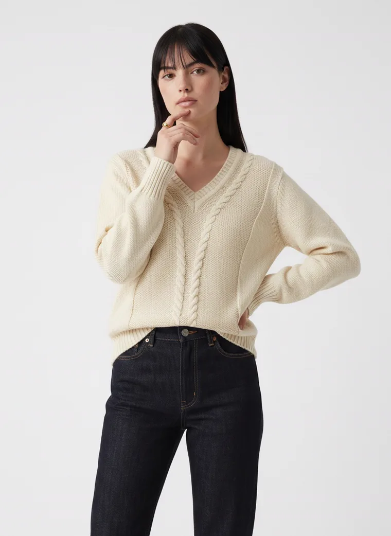 A cream cable knit v-neck jumper with black straight jeans on pale grey. Cable Knit V-Neck in Cream, frame 3 of 5