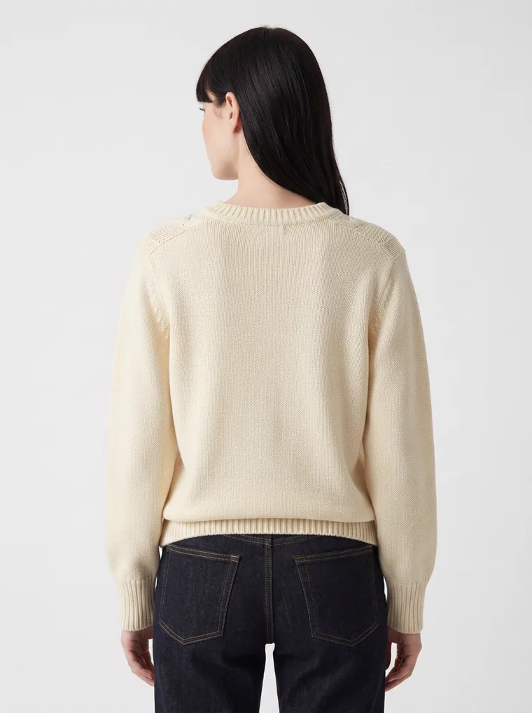 A cream cable knit v-neck jumper with black straight jeans on pale grey. Cable Knit V-Neck in Cream, frame 4 of 5